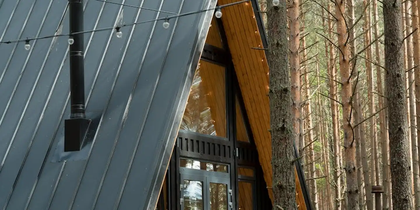 A-frame house with a metal-panel roof in a forest setting, featuring a large glass entry and timber interior framing.