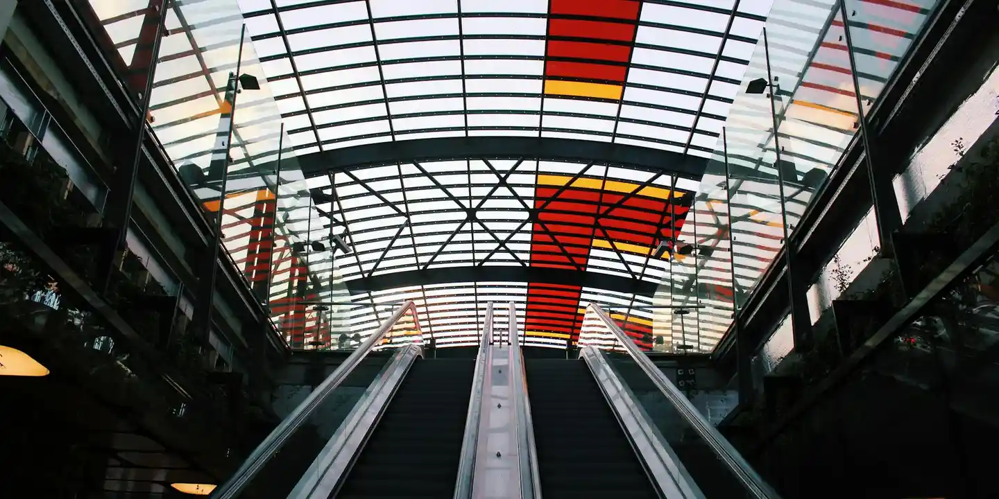 Interior view of a modern glass-roof atrium with a grid of metal framing and skylight panels, seen from a lower level with escalators.