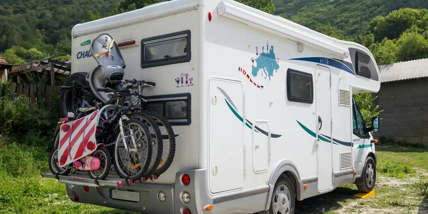 White motorhome with a rear-mounted bicycle rack parked in a rural area with trees and hills in the background.