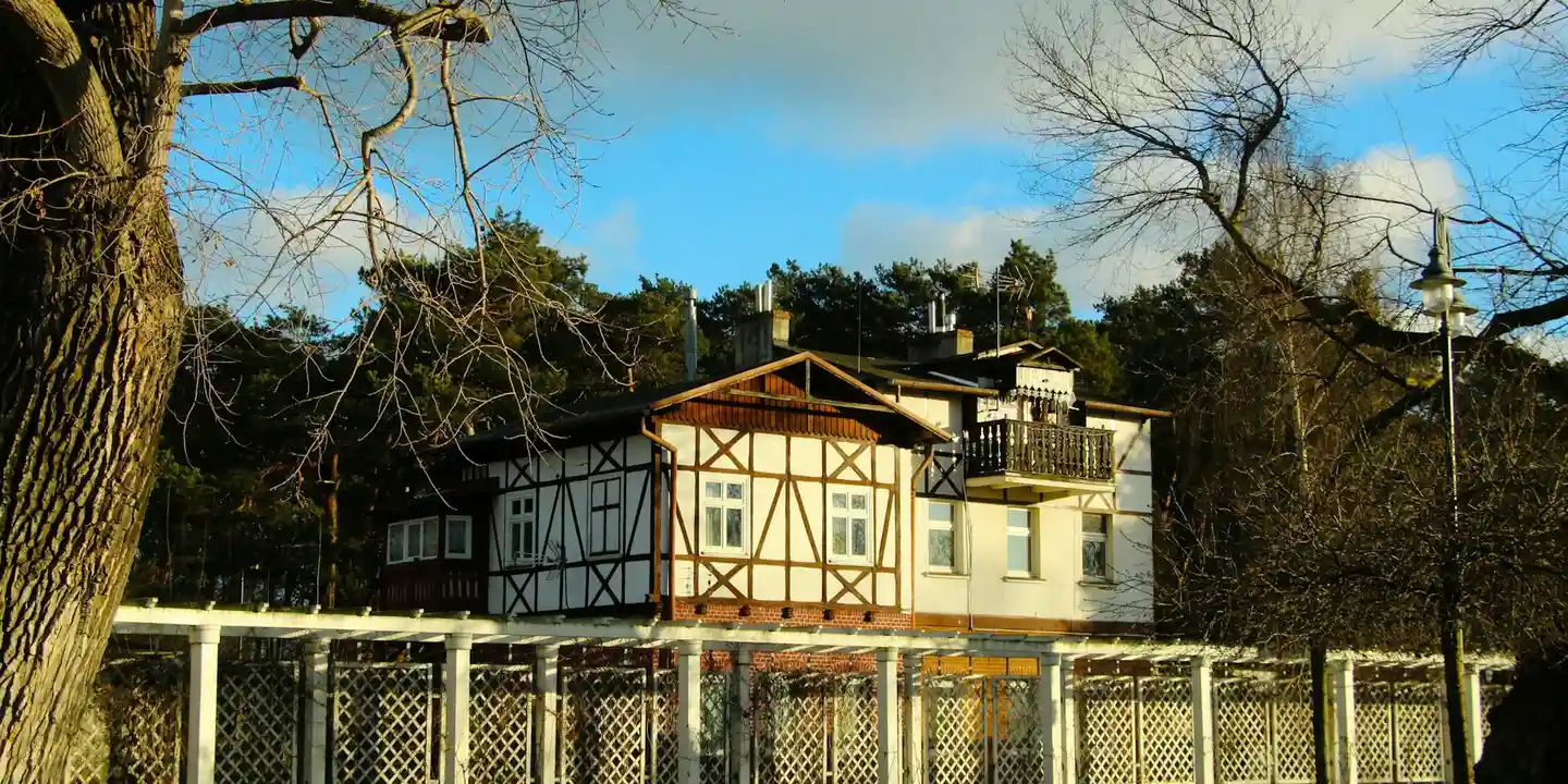 Two-story house with Tudor-style framing and a visible chimney; a chain-link fence is in the foreground beneath a blue sky.