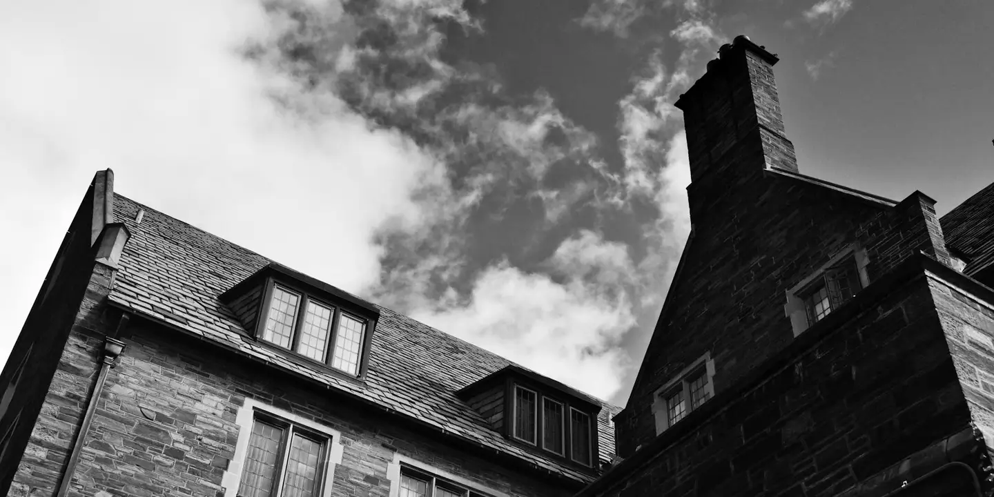 Black-and-white photograph of a house with a tall chimney on the roof, dormer windows on the left, and a steep pitched roof under a cloudy sky.