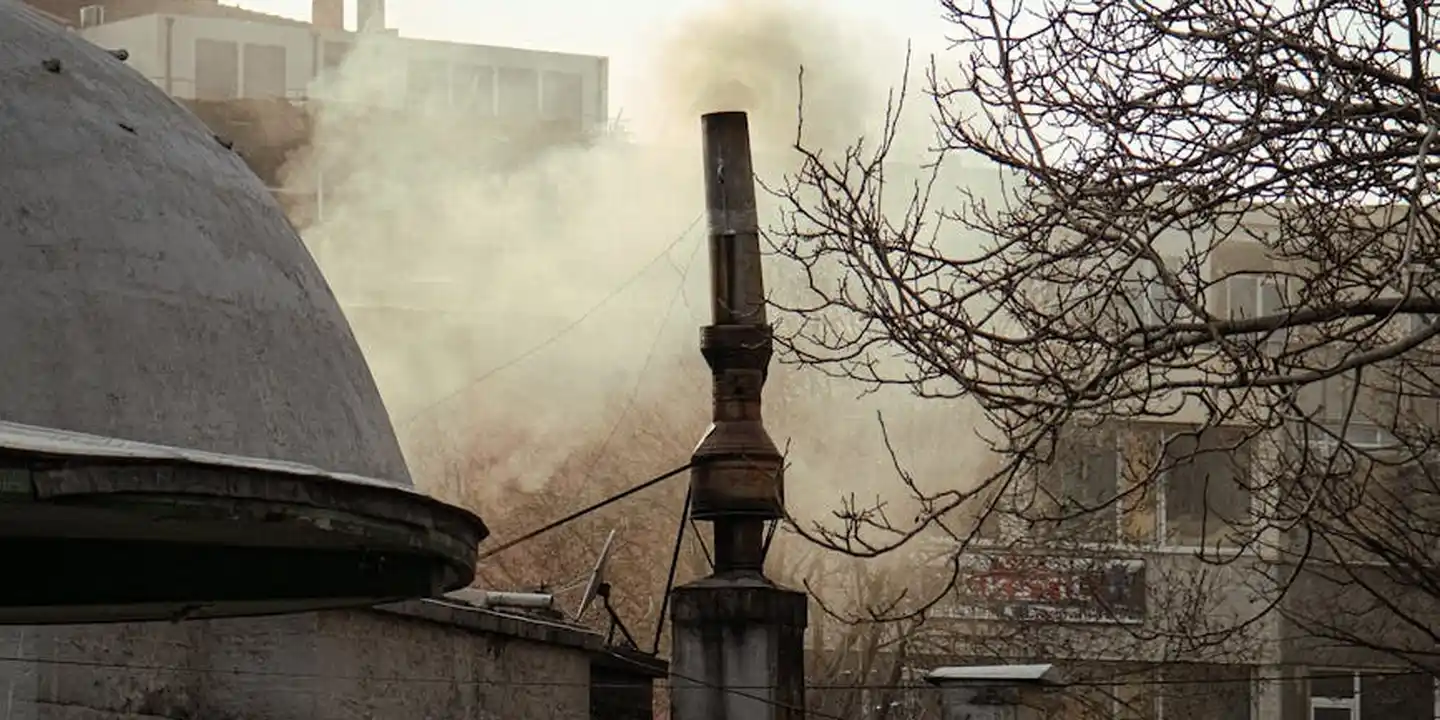 Smoke rising from a metal chimney stack on a roof with a bare tree nearby.