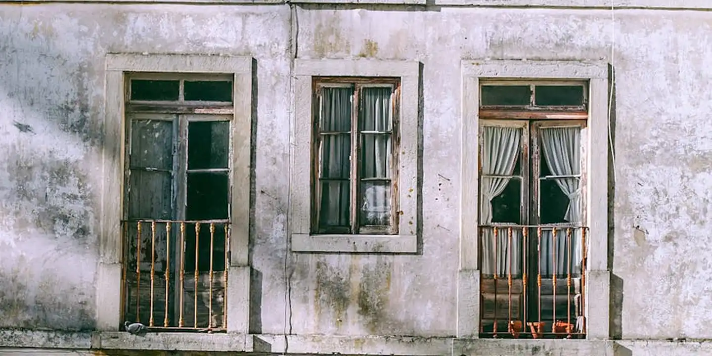 Weathered exterior wall with three window openings, one with a small balcony railing and tattered curtains, showing wear that highlights the need for proper roof and chimney maintenance to prevent wildlife entry.