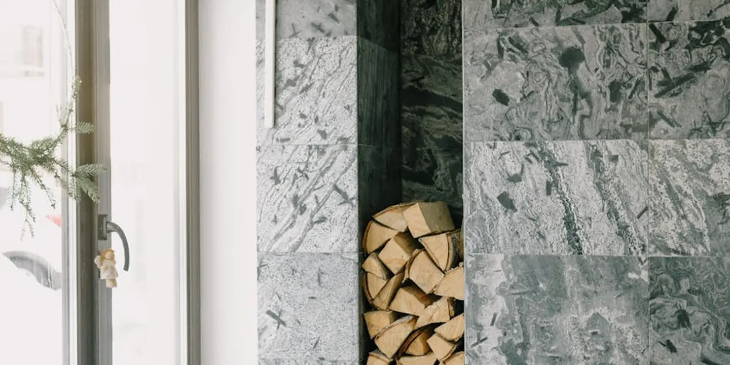 Indoor scene showing marble-patterned walls, a doorway to the left, a dark vertical column in the center, and a stack of firewood stored in a cabinet on the right.
