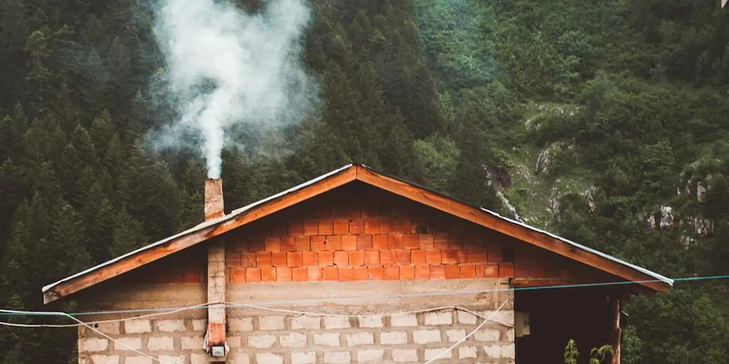Chimney on a small wooden house emitting smoke with a forested backdrop, illustrating chimney waterproofing considerations.