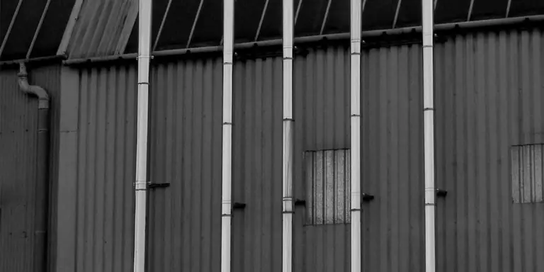 Vertical vent pipes attached to a corrugated metal exterior wall, illustrating exterior venting for a stove without a chimney.