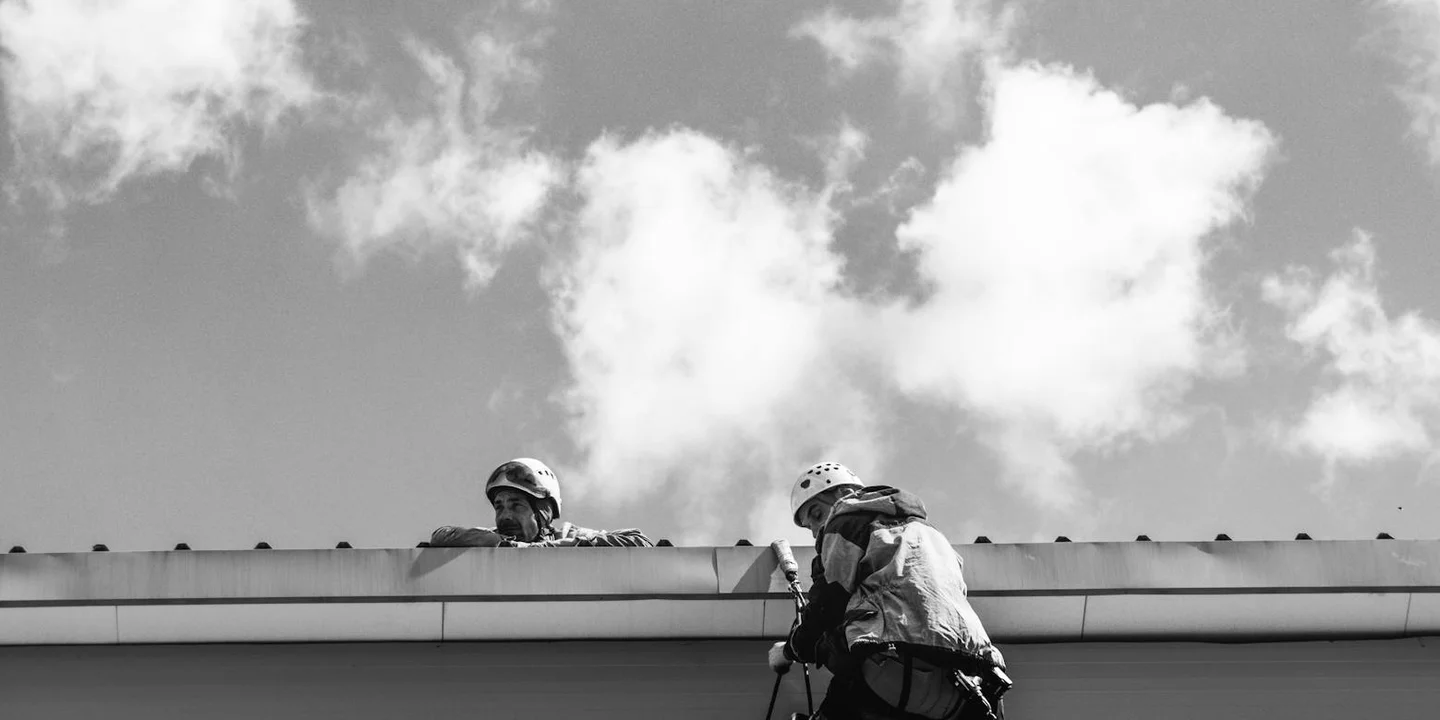 Two roof inspectors in safety gear evaluate a flat commercial roof from the edge against a cloudy sky.