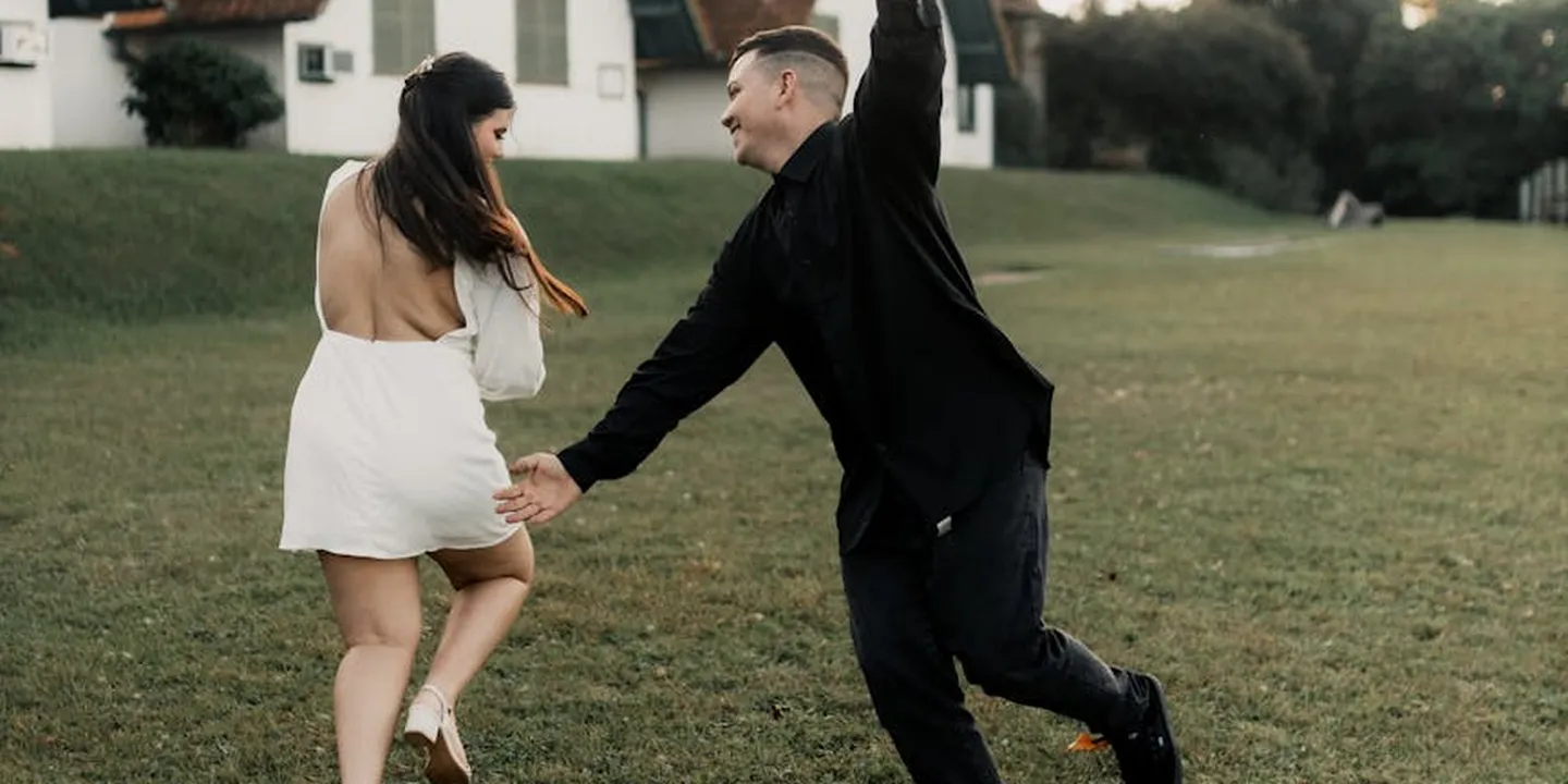 Two people outdoors near a house: a man in dark clothing raises one arm while a woman in a white dress walks away in the grassy yard.