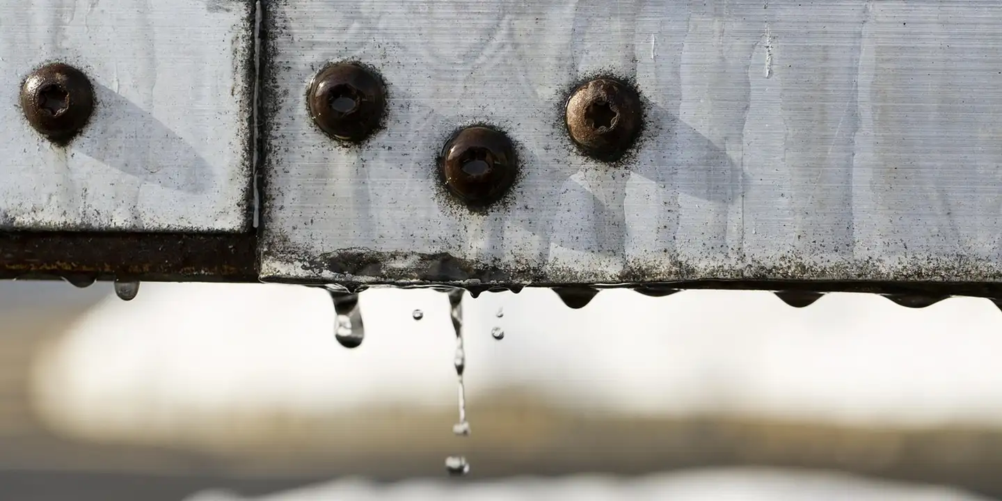 Close-up view of a metal roof drip edge along the eaves, with water droplets hanging from the edge and exposed screws.
