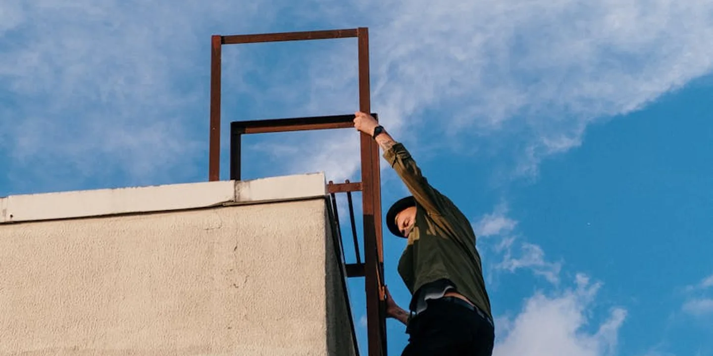 A construction worker on a roof adjusting metal drip-edge components along the eave, with a ladder nearby and a blue sky.
