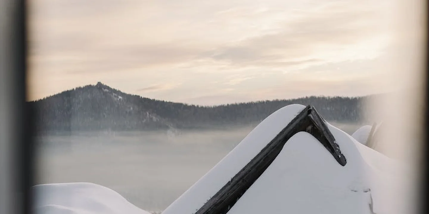 Snow-covered roof with a dark tarp over the ridge, viewed through a window frame, illustrating emergency tarping work.