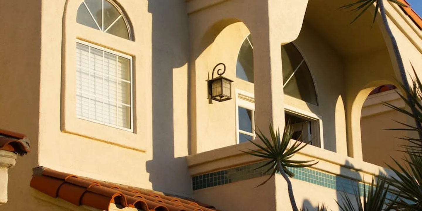 Front view of a stucco house with arched windows and a Spanish-style clay tile roof edge, with a small palm plant nearby.
