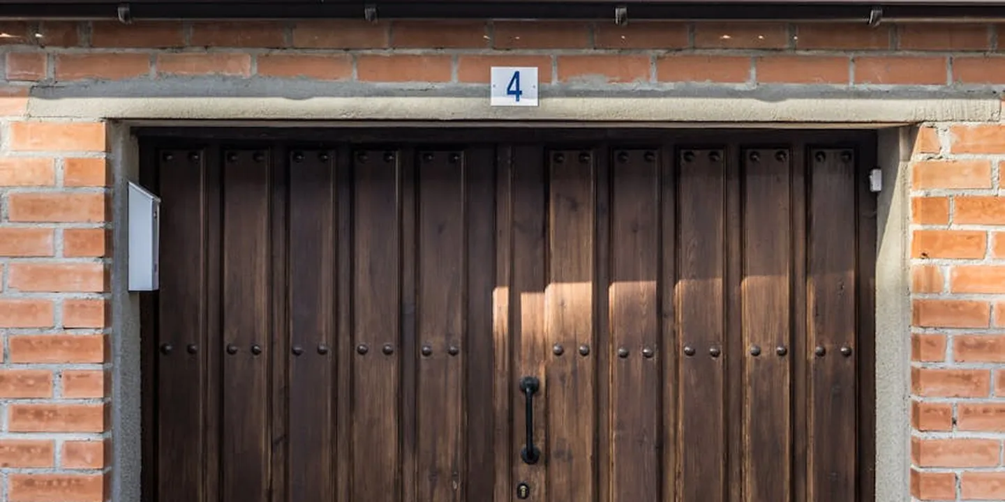 A dark brown wooden garage door set in a brick exterior, with a small blue-and-white house number plaque above and sunlight creating a rectangular highlight on the door.
