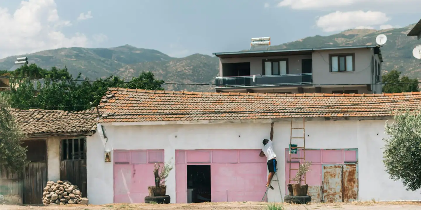 A person on a ladder repairing the roof of a small garage with pink doors, against a hillside backdrop.