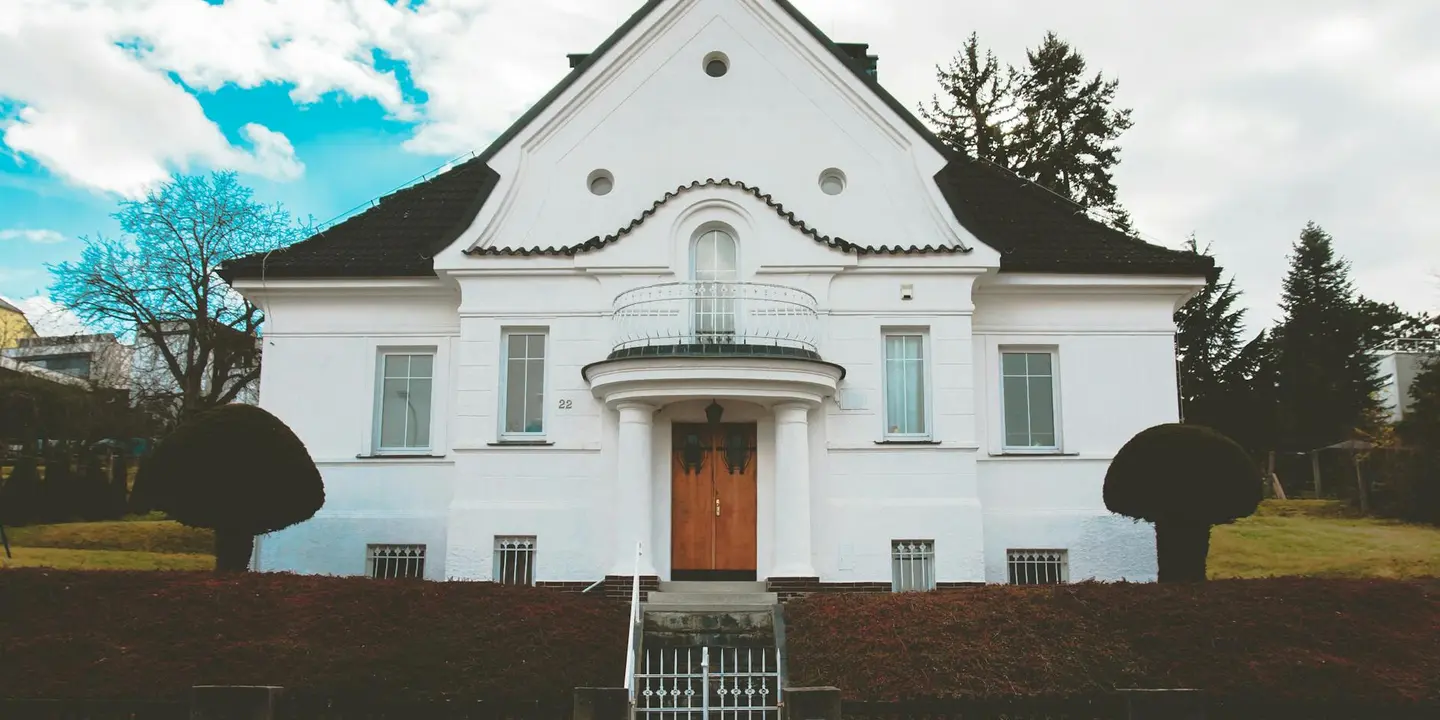 White two-story house with a central arched doorway, symmetrical windows, and neatly trimmed spherical shrubs in a tidy yard under a bright sky.