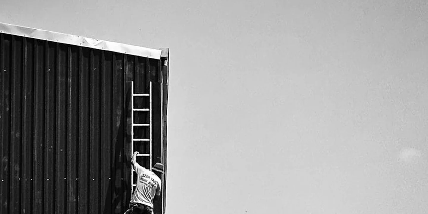 Black-and-white image of a person climbing a ladder against a house to access the roof for inspection