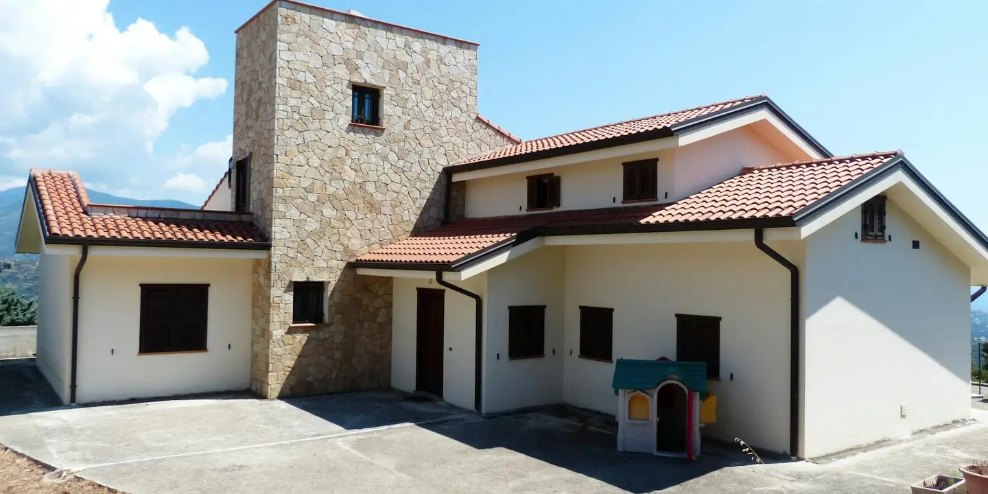 A modern house featuring a tall stone chimney stack, white stucco walls, and a red clay tile roof under a clear blue sky.