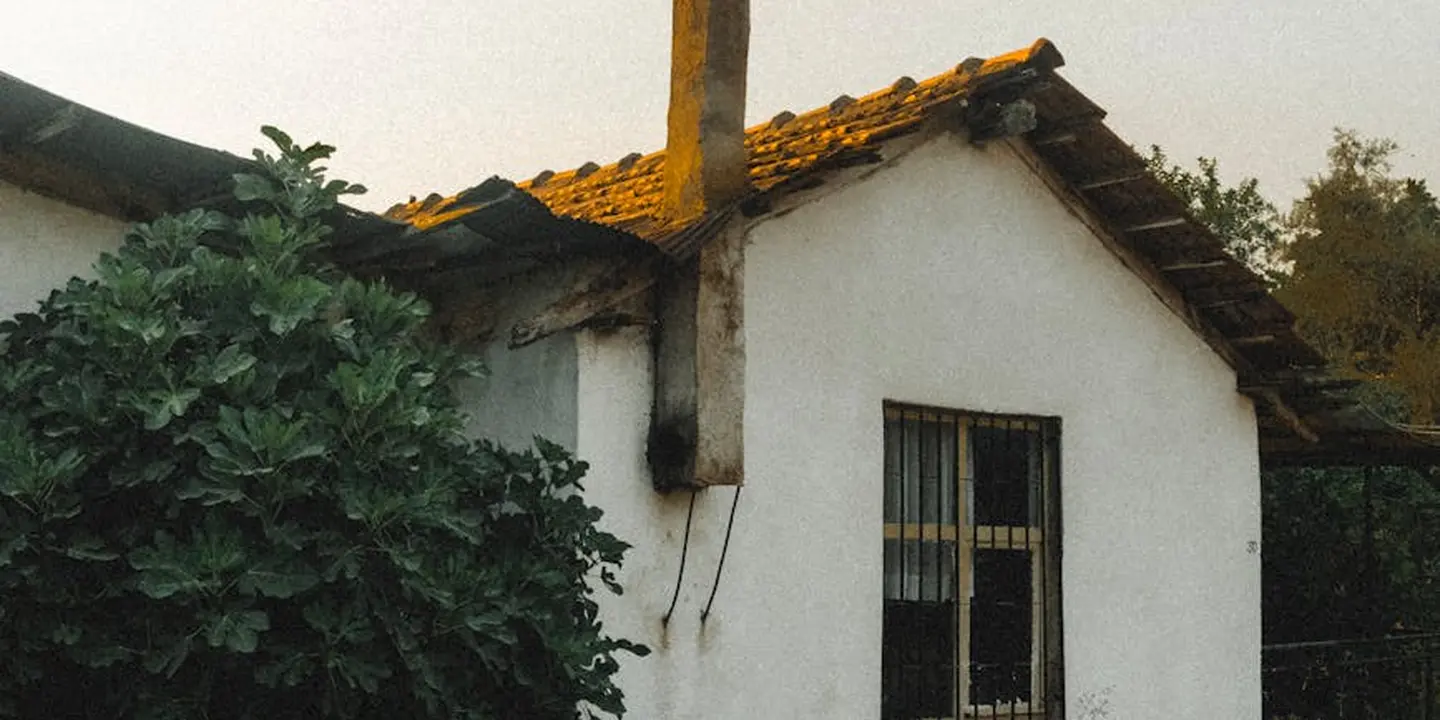 Small white house with a tiled roof and a vertical chimney-like vent protruding from the roof, with greenery in the foreground.