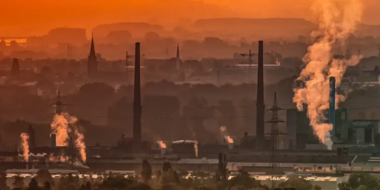 Industrial skyline with smokestacks emitting smoke against an orange sunset.