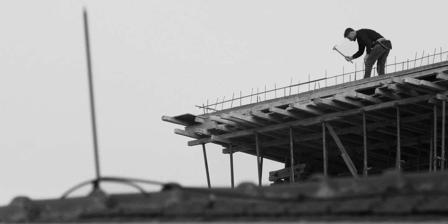 A construction worker stands on a rooftop scaffold, examining the roof framing.