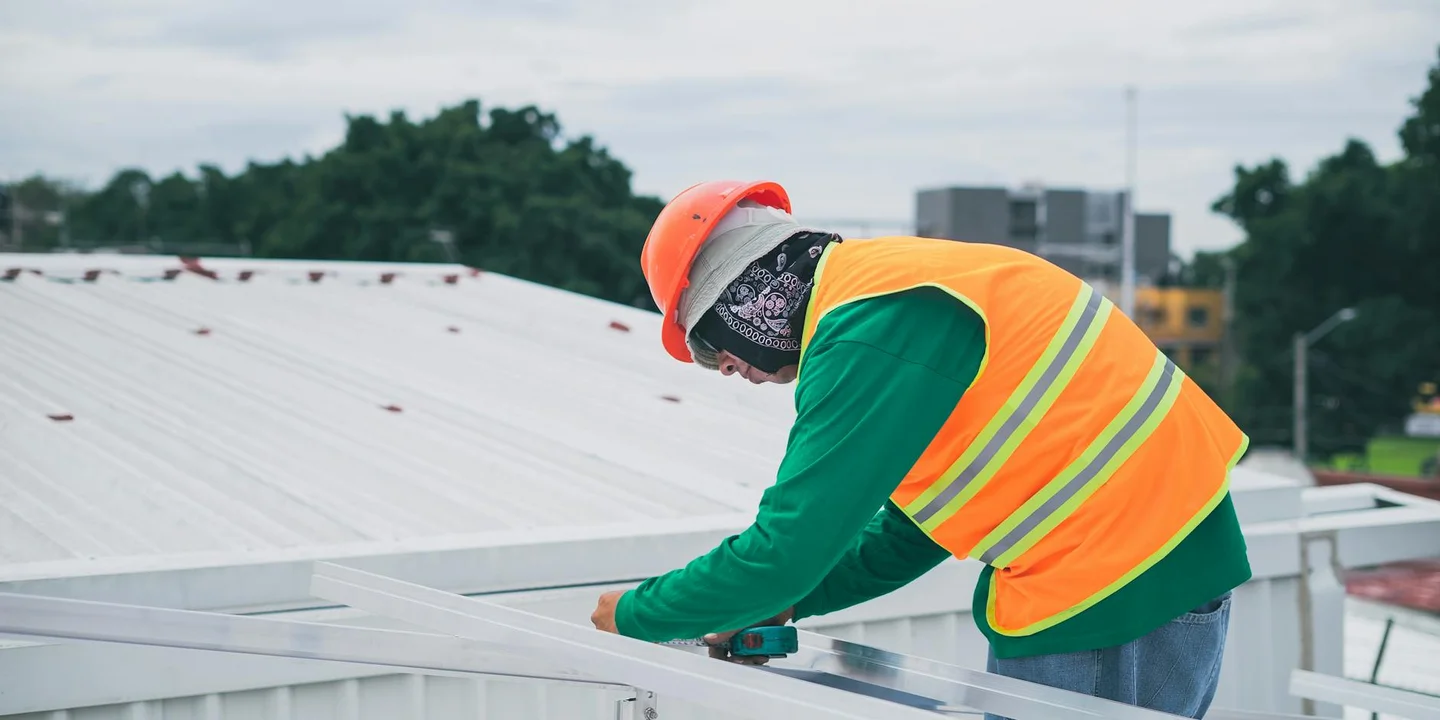 Worker on a metal roof wearing a high-visibility vest and hard hat, preparing roofing materials and tools to manage dust and fumes and establish a safe worksite.