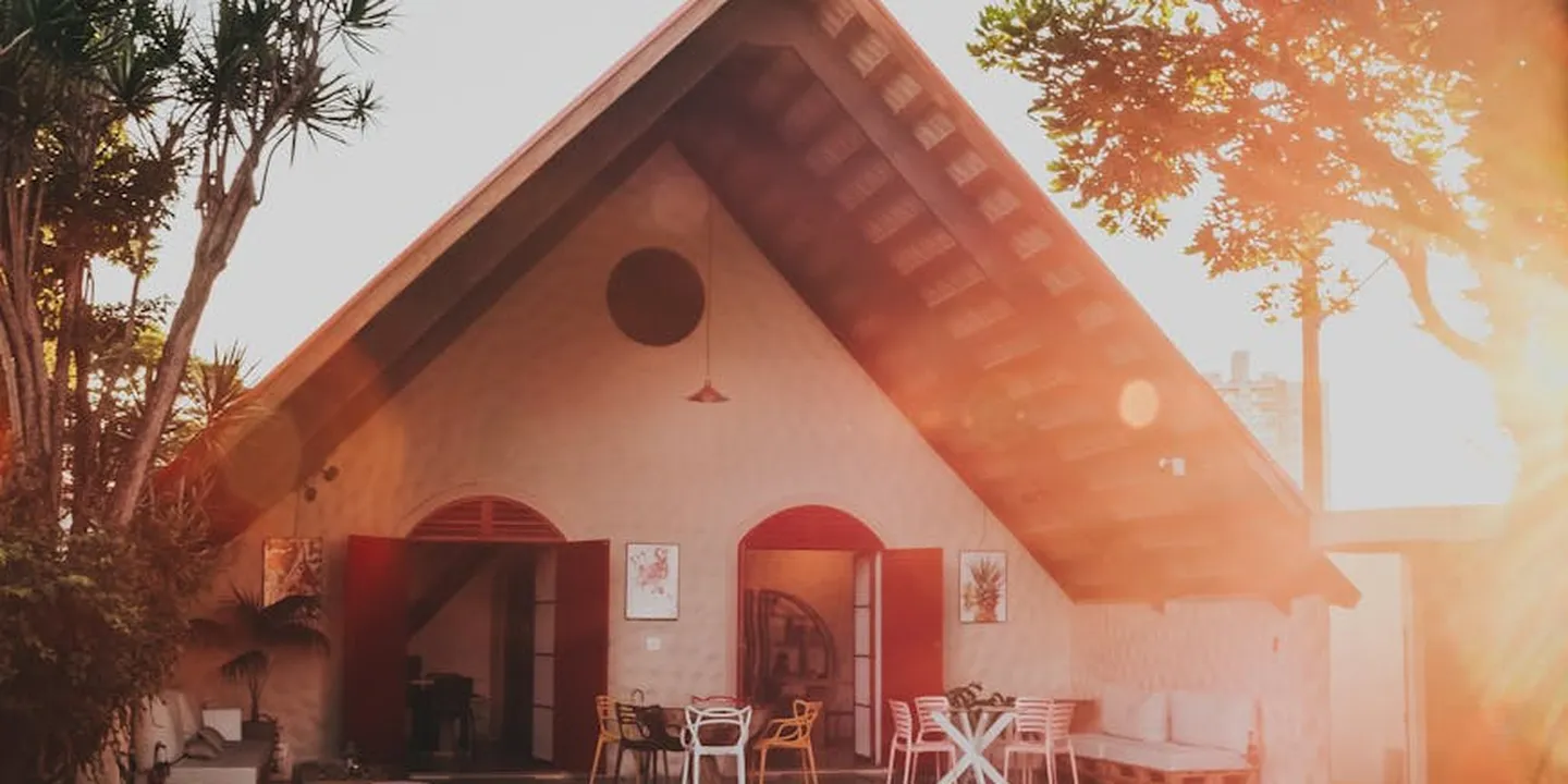 A small house with a steep gable roof and an inviting shaded porch, featuring outdoor seating and warm sunlight streaming from the right.