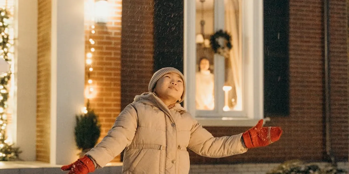 Person in a winter jacket and red gloves stands beside a house with string lights, illustrating planning the outdoor lighting layout from ground level.