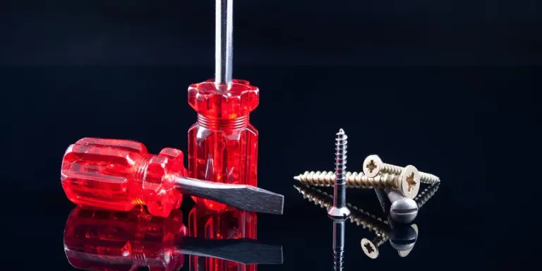 Close-up of red-handled screwdriver and metal screws with bits on a reflective surface, illustrating roof fasteners.