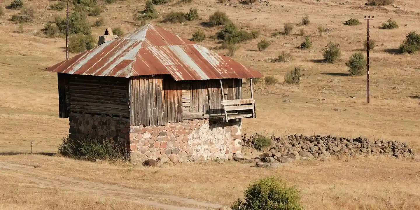 A small wooden shed with a weathered, rusted corrugated metal roof in a dry, grassy field