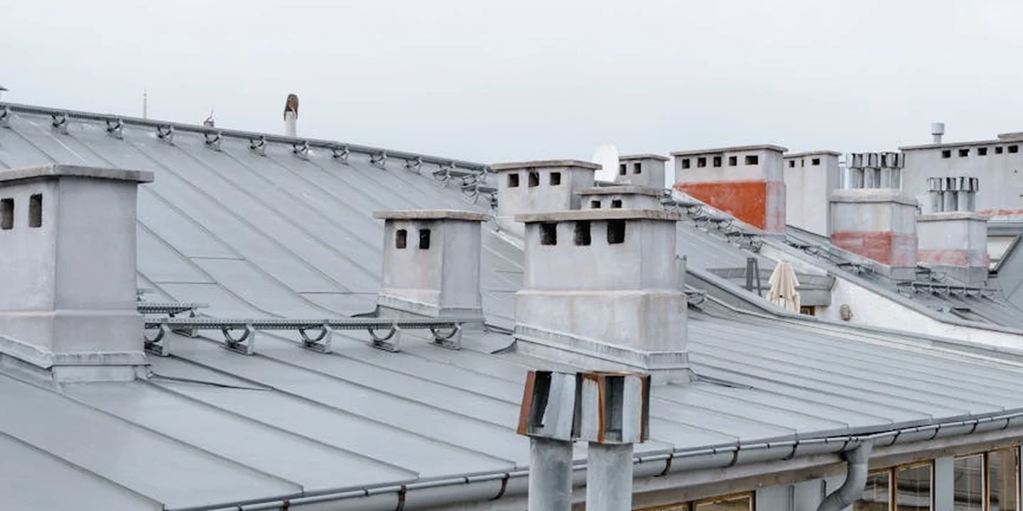 Row of metal roofs with chimneys and vents under a grey cloudy sky, illustrating durable, low-maintenance roofing material.