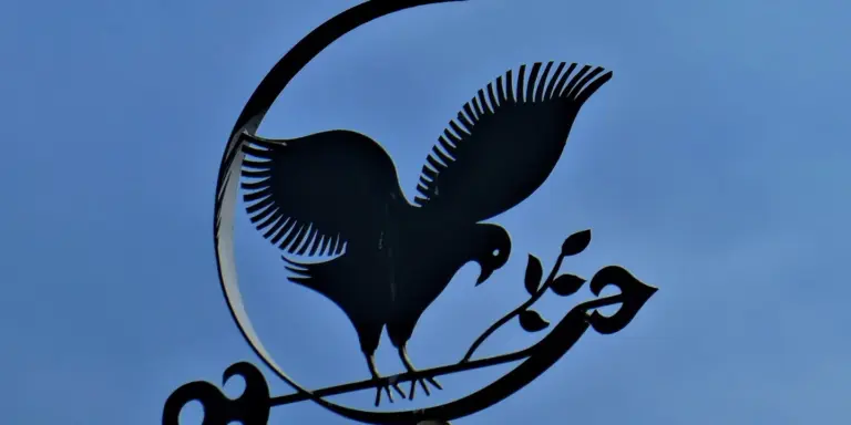 Silhouette of a decorative black metal bird sculpture perched on branches against a blue sky, representing metal craftsmanship related to roofing.