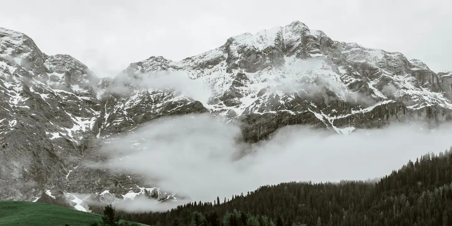 Snow-covered mountain ridge with clouds and a forested slope beneath