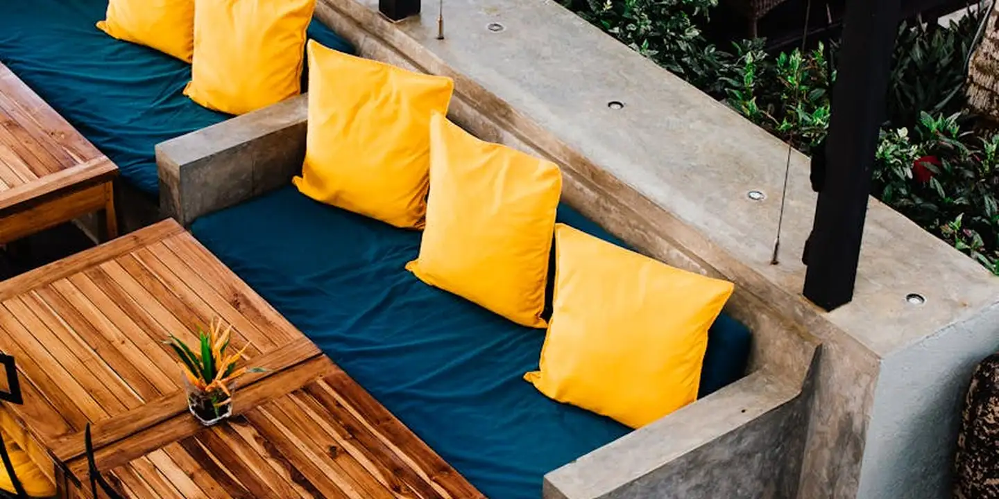 Outdoor patio seating area with a built-in bench, blue cushions, and bright yellow pillows along a concrete edge under a pergola, illustrating cozy finishing touches for outdoor living spaces.