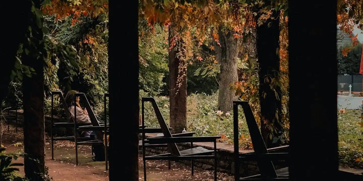 Outdoor pergola with vertical posts and a slanted roof, set in a shaded area with autumn leaves; a person sits on a bench beneath the structure.