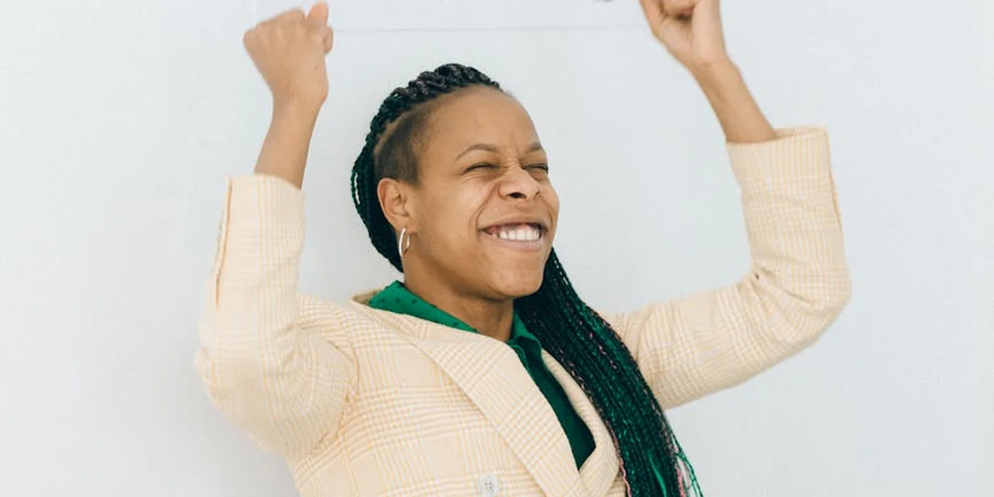 Smiling woman with braided hair raises her fists in celebration against a light, neutral background