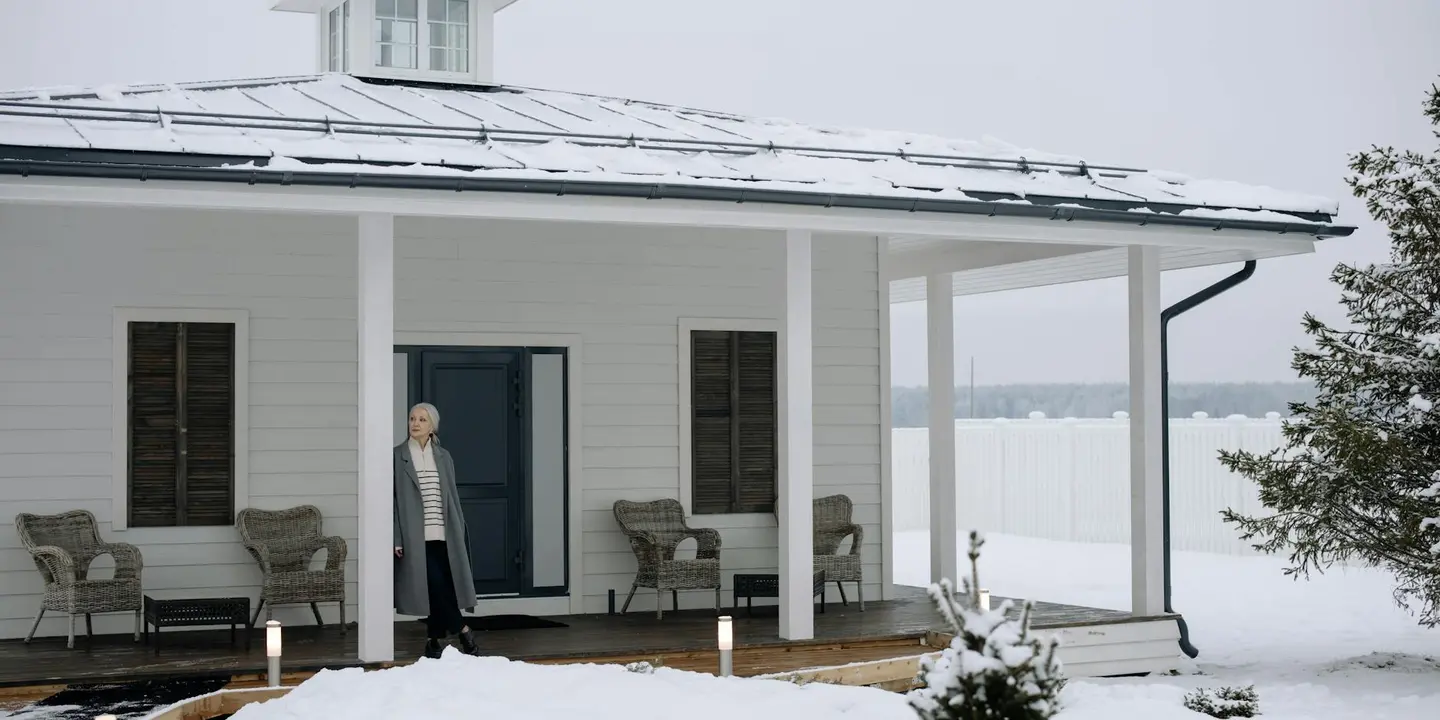 A man stands on a snow-covered porch of a white house, with outdoor seating and a wintry landscape.