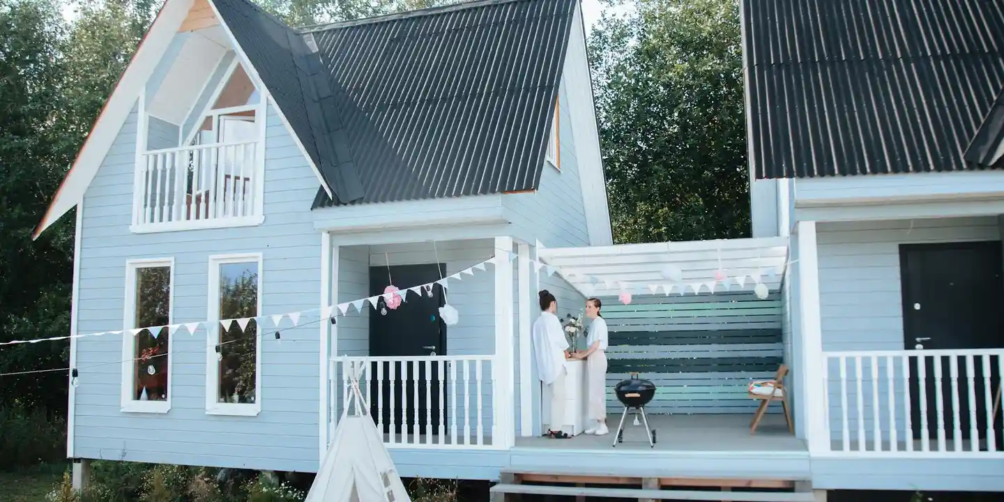Blue house with white porch railing and a covered patio roof extension; string lights across the porch; two people stand on the porch near a small grill.