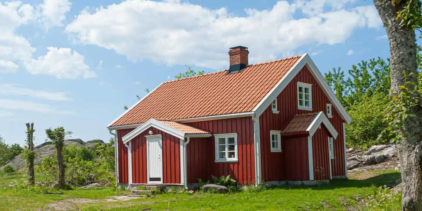 Small red wooden house with white trim and a red-tiled roof, set in a green lawn under a partly cloudy sky.