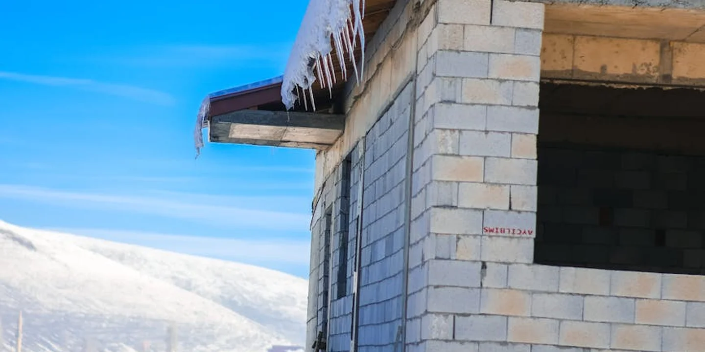 Partially built residential wall made of concrete blocks with an opening, icicles hanging from the eave, and a snow-covered roof against a blue sky.