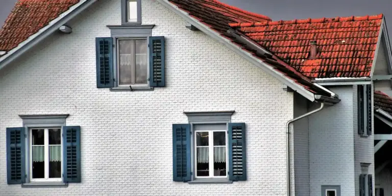 White brick house with blue window shutters and a red tile roof featuring a dormer window, illustrating a traditional pitched residential roof