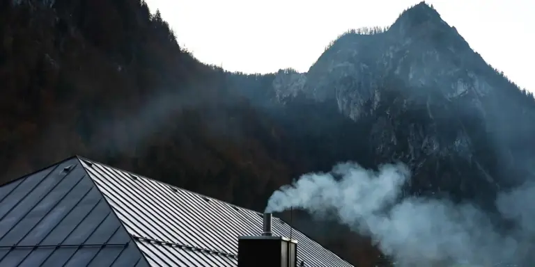 A residential roof with a metal seam roof and a chimney emitting smoke, with mountains in the background.
