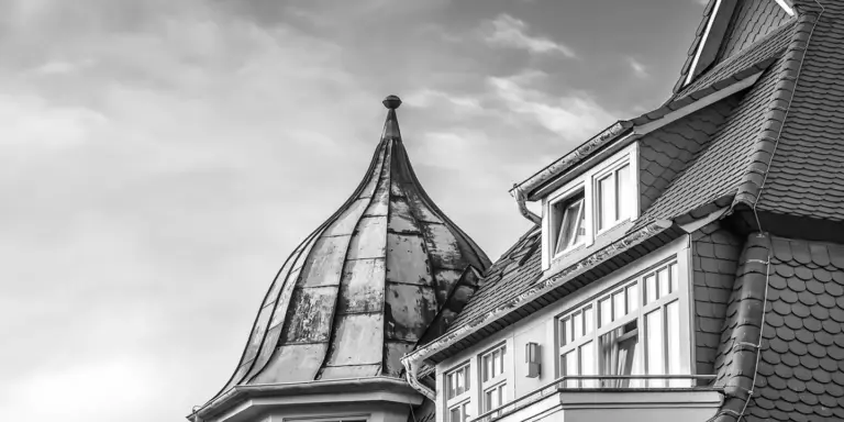 Black-and-white photo of a residential roof with tile shingles and a turret against a cloudy sky.