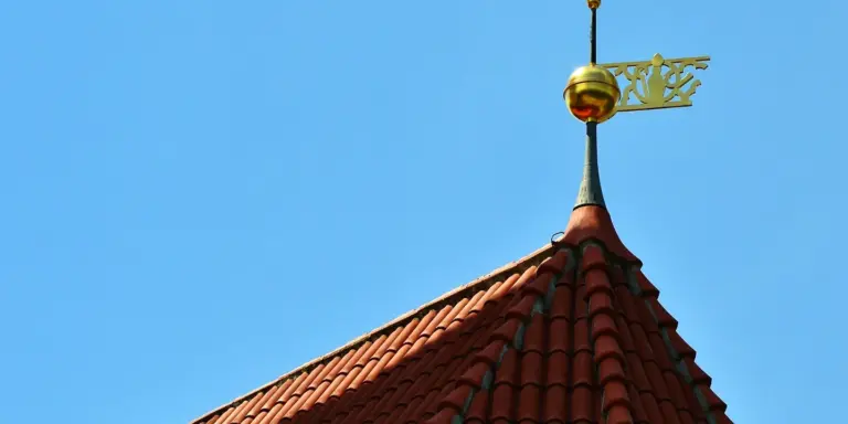Residential roof with red clay tiles and a weather vane against a bright blue sky.