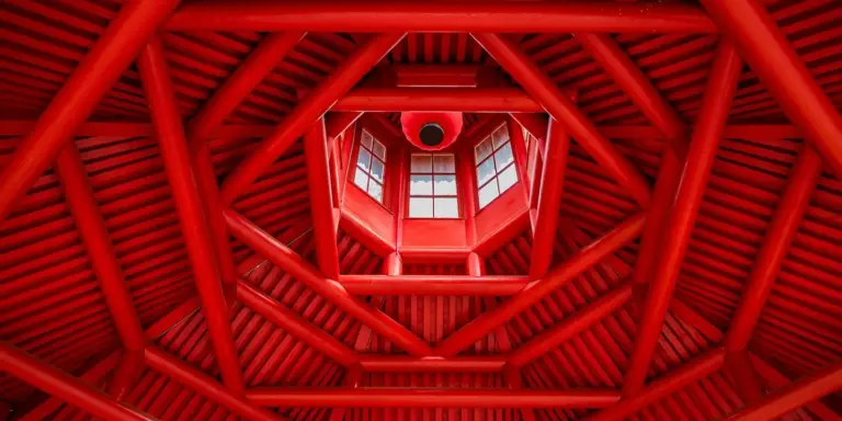 Inside view of a red-painted wooden roof structure with exposed trusses and a central window, showing the complex framing before roof sheathing is applied.