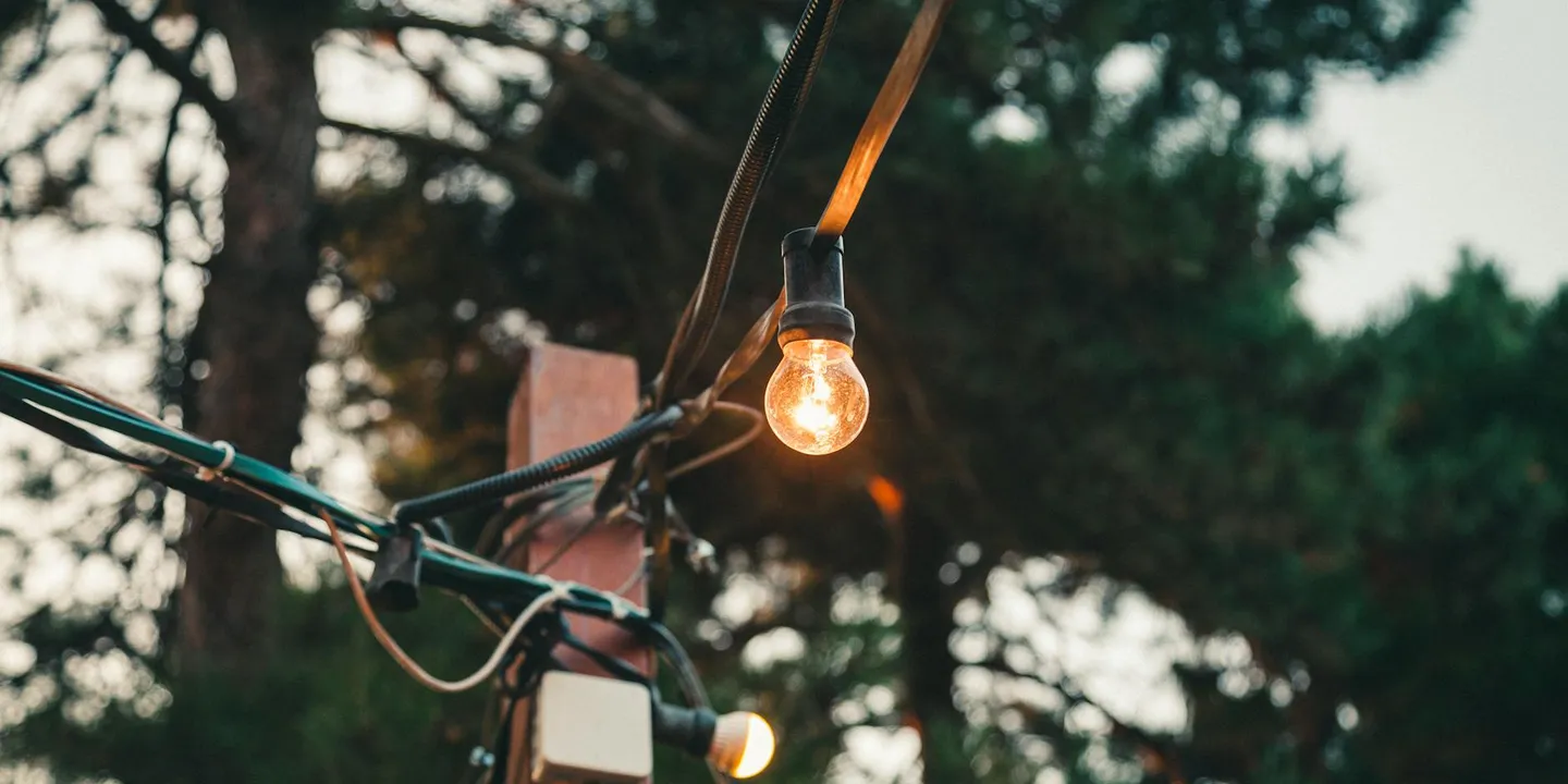 Outdoor string lights with a single glowing bulb hanging from a cord, wires secured along a railing or roof edge in an outdoor setting.