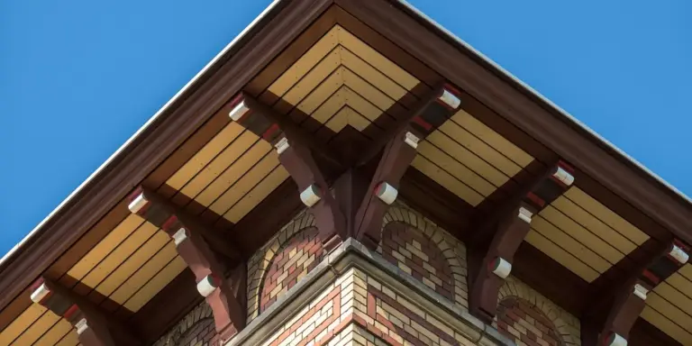 Close-up of a roof edge with exposed wooden decking boards and supporting trusses against a clear blue sky.