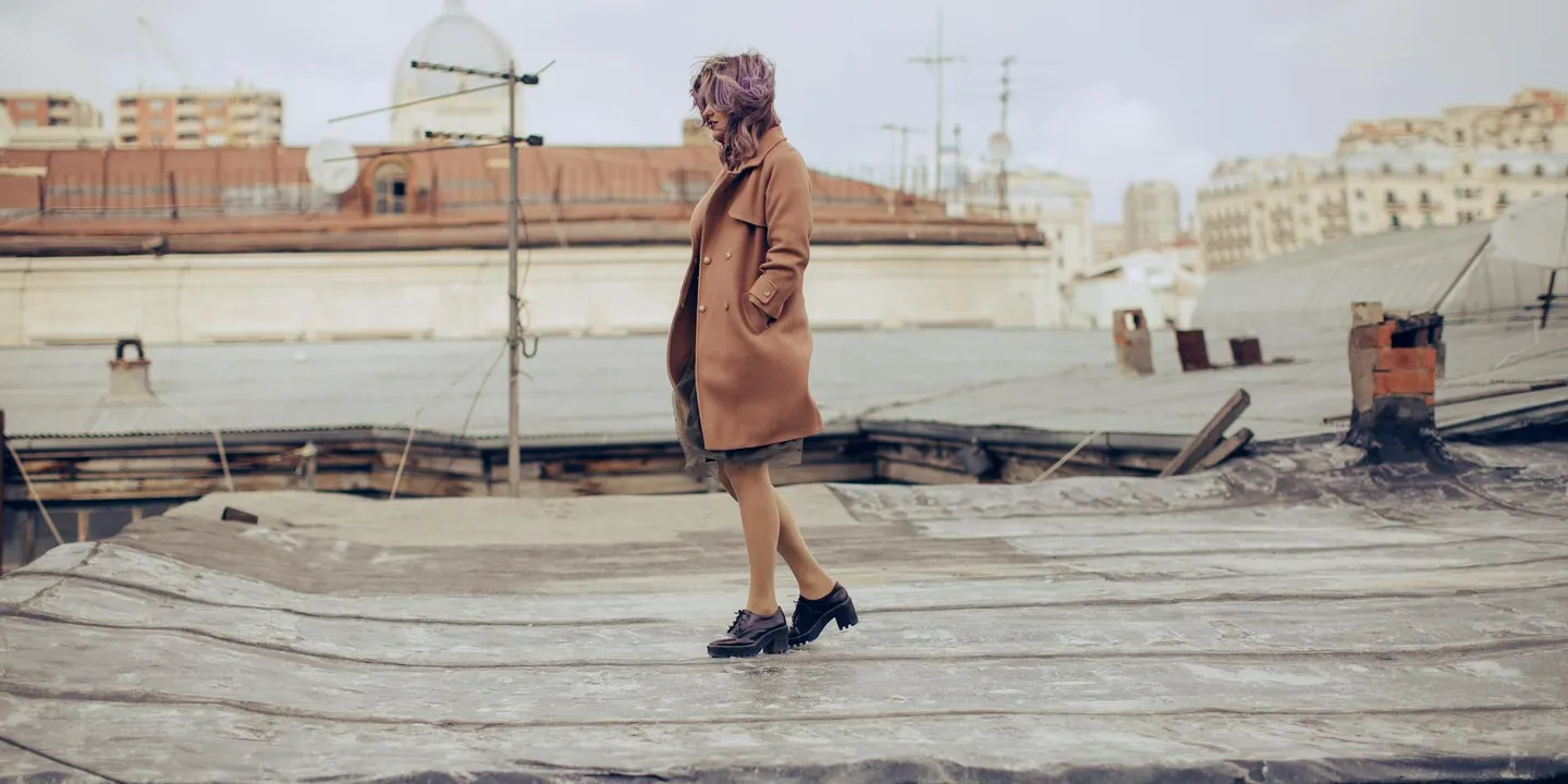 A woman in a brown coat walks on a damaged rooftop with scattered debris and collapsed sections, illustrating roof damage and insurance claim challenges.