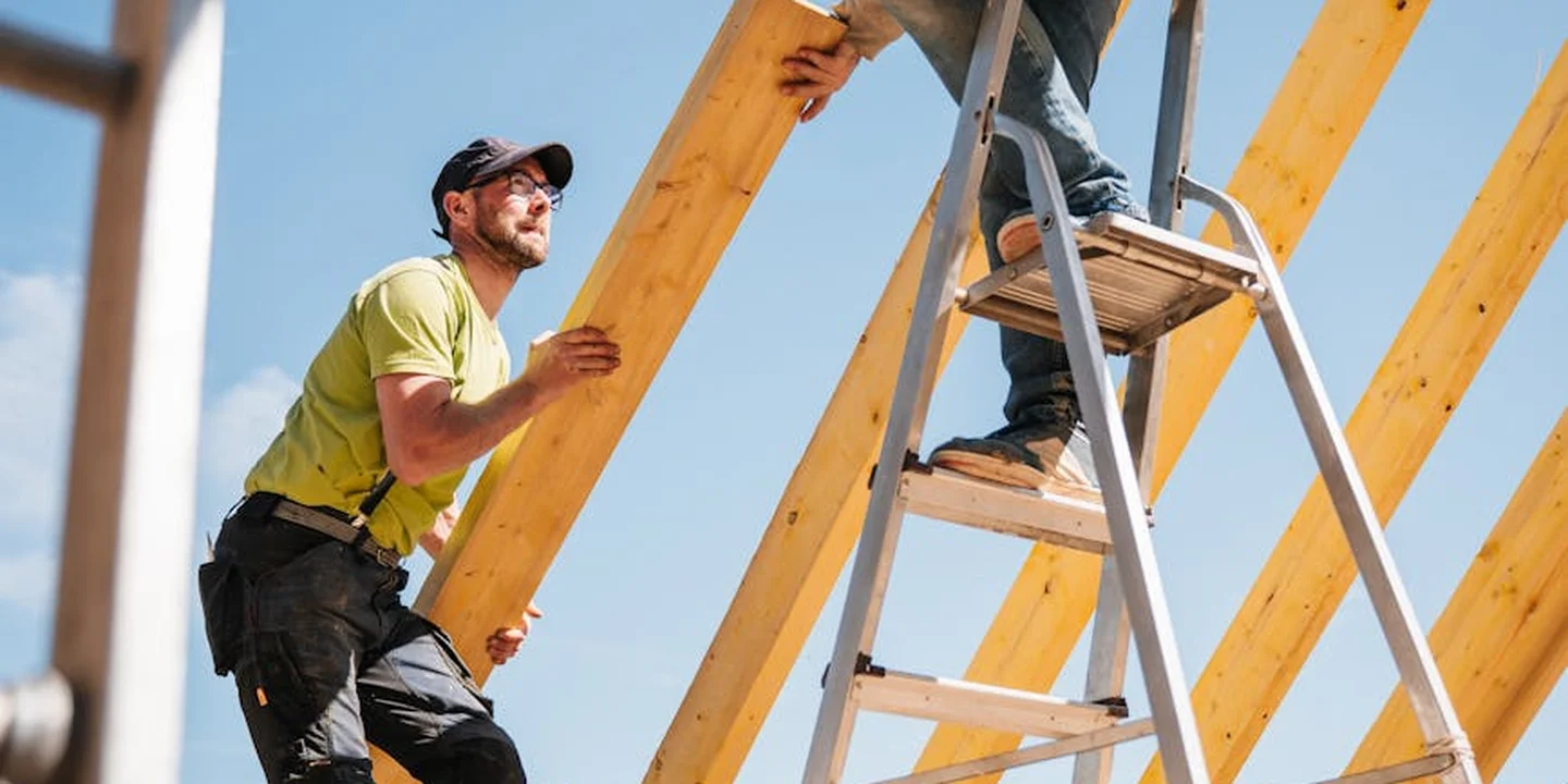 Two construction workers on a roof frame; one on a ladder and another holding a wooden beam, illustrating ladder use for roof access.