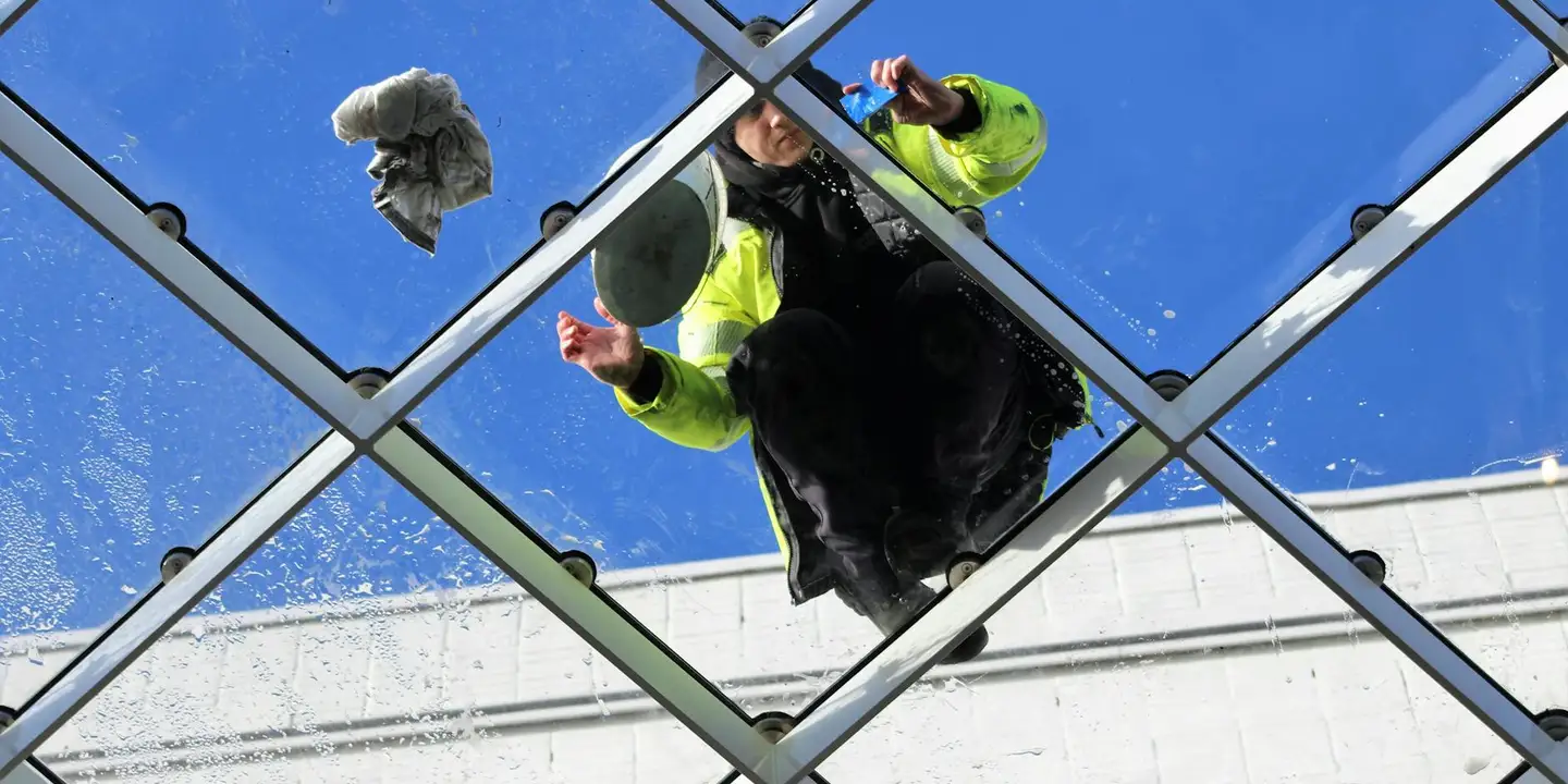 Two workers wearing high-visibility jackets on a glass skylight, looking down and inspecting or cleaning the roof from above against a blue sky.