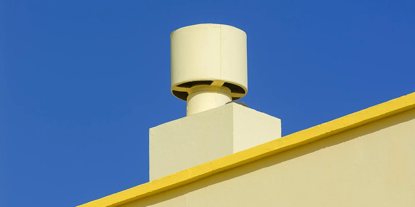 A round vent cap on a chimney-like vent protrudes from a pale structure with a yellow edge, set against a clear blue sky.